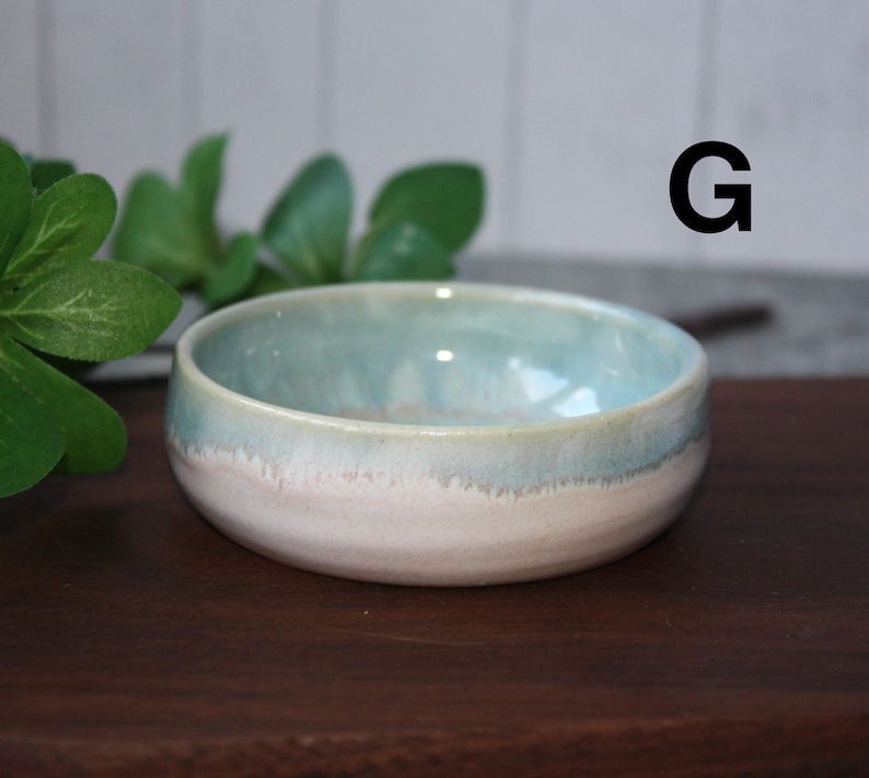May include: A small, handmade ceramic bowl with a light blue and white glaze. The bowl has a rounded shape and a smooth, glossy finish. The letter "G" is visible in the background. The bowl is likely used for serving food or as a decorative item.