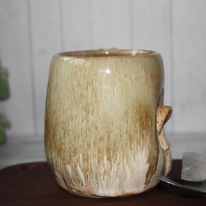 Pode incluir: Uma caneca de cer&acirc;mica artesanal com uma forma org&acirc;nica &uacute;nica. A caneca apresenta um esmalte bege com riscas castanhas, criando uma apar&ecirc;ncia texturizada e terrosa. Uma pega esculpida adiciona ao design art&iacute;stico da caneca. Perfeita para caf&eacute; ou ch&aacute;.