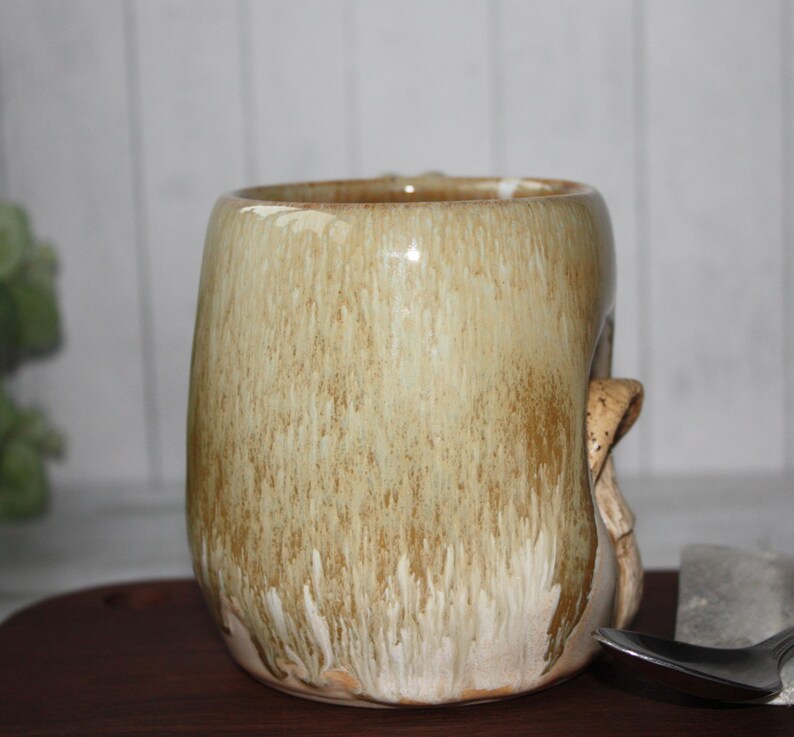 Pode incluir: Uma caneca de cer&acirc;mica artesanal com uma forma org&acirc;nica &uacute;nica. A caneca apresenta um esmalte bege com riscas castanhas, criando uma apar&ecirc;ncia texturizada e terrosa. Uma pega esculpida adiciona ao design art&iacute;stico da caneca. Perfeita para caf&eacute; ou ch&aacute;.