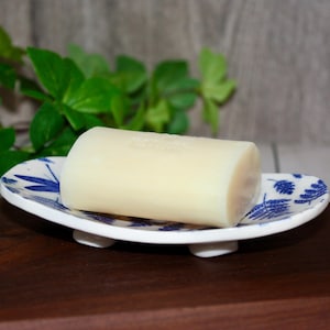 May include: A pale bar of soap sits on a white soap dish with a blue floral pattern. The soap dish has three small white feet. The background features green leaves and a wooden surface.