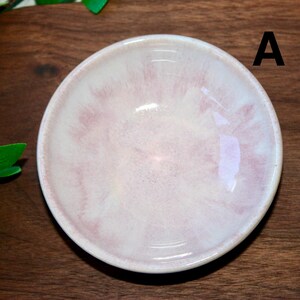 May include: A small, round ceramic bowl with a pale pink and white marbled design. The bowl has a glossy finish and a slightly raised rim. The letter "A" is visible on the right side of the bowl. The bowl is likely used for serving small portions of food or as a decorative item.