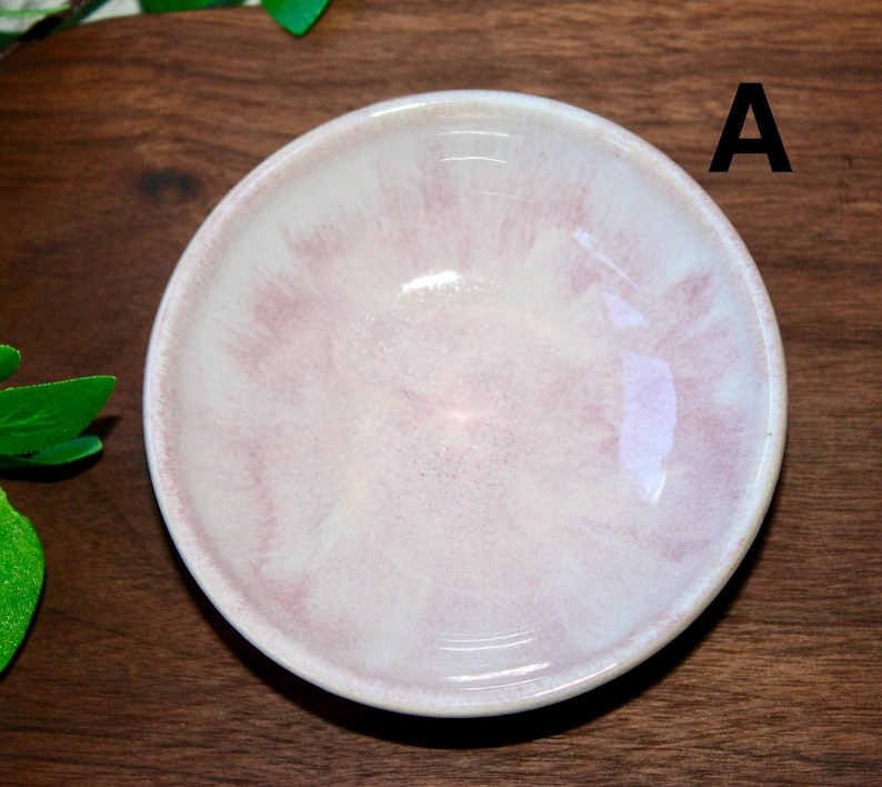 May include: A small, round ceramic bowl with a pale pink and white marbled design. The bowl has a glossy finish and a slightly raised rim. The letter "A" is visible on the right side of the bowl. The bowl is likely used for serving small portions of food or as a decorative item.