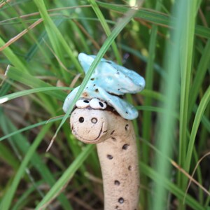 May include: A ceramic garden stake featuring a tan, cylindrical worm-like figure with a smiling face, two white eyes, and a light blue, textured hat-like accessory resting on its head, decorated with small dark brown spots along its body.