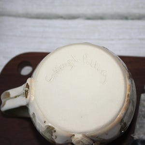 Pode incluir: Uma caneca de cer&acirc;mica artesanal com interior creme e exterior texturizado branco sujo com detalhes em castanho e verde. A base da caneca apresenta a inscri&ccedil;&atilde;o "Collingwood Pottery". A caneca tem uma pega e est&aacute; colocada numa superf&iacute;cie de madeira.