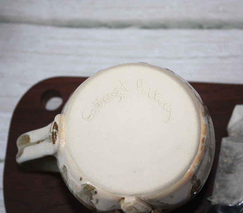Pode incluir: Uma caneca de cer&acirc;mica artesanal com interior creme e exterior texturizado branco sujo com detalhes em castanho e verde. A base da caneca apresenta a inscri&ccedil;&atilde;o "Collingwood Pottery". A caneca tem uma pega e est&aacute; colocada numa superf&iacute;cie de madeira.