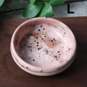 May include: A small, round ceramic bowl with a speckled pink interior and a cream-colored rim. The bowl has a glossy finish and is decorated with small black spots. The bowl is likely handmade and suitable for serving small portions.