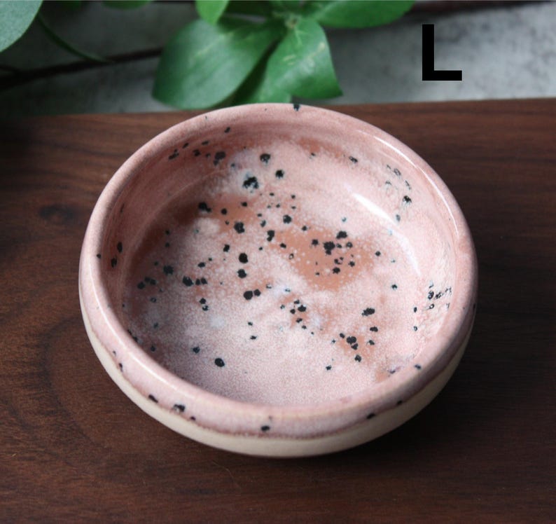 May include: A small, round ceramic bowl with a speckled pink interior and a cream-colored rim. The bowl has a glossy finish and is decorated with small black spots. The bowl is likely handmade and suitable for serving small portions.