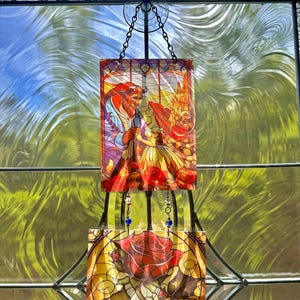 Op de afbeelding: Gebrandschilderd glaskunst met een scène met het Beest en Belle, en een rode roos. Het kunstwerk wordt opgehangen met een ketting en is omlijst met een zwarte metalen structuur. De kleuren zijn levendig, met rood, geel en blauw.
