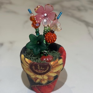 May include: A miniature decorative flower pot with a fabric exterior featuring a sunflower design. The pot contains artificial flowers with pink, orange, and clear petals, green leaves, and beaded accents. The pot is filled with green moss.