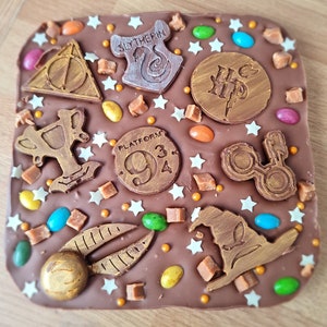 May include: A chocolate bar with a Harry Potter theme. The bar is decorated with various Harry Potter symbols, including the Hogwarts crest, the Golden Snitch, and the Platform 9 3/4 sign. The bar is also decorated with colourful sprinkles and candy.