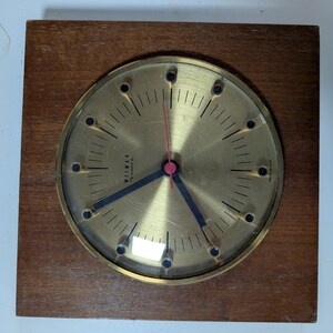 May include: A vintage Weimar quartz clock with a gold-colored face and black hands. The clock is set in a square, wooden frame. The clock face has black markers and a red second hand. The clock face has the text "WEIMAR QUARTZ" printed on it.