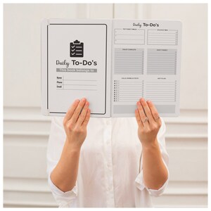 May include: A white daily planner with a black checklist icon and the text "Daily To-Do's". The planner has sections for "Top Three Things", "Must Complete", "Don't Forget", "Calls, Emails & Texts", and "Not To Do".