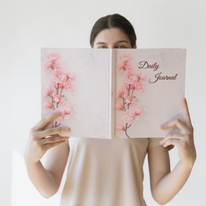 以下が含まれることがあります： 女性が、表紙に「Daily Journal」と印刷されたピンクと白のジャーナルを開いて持っています。ジャーナルは、ピンクの花が咲いた花柄のデザインです。