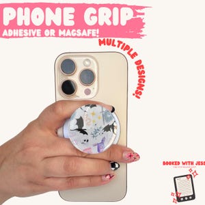 May include: A white phone grip with a circular design featuring bats, stars, and other whimsical elements. The phone grip is attached to a gold-colored phone. Text on the image reads "PHONE GRIP" and "ADHESIVE OR MAGSAFE!" and "MULTIPLE DESIGNS!"