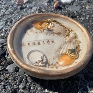 May include: A decorative ceramic dish with a gold rim and the word "seaside" spelled out in small letters. The dish contains seashells, sand, and jewelry, including a gold necklace and a silver ring.