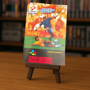 Può includere: Una scatola di gioco per Super Nintendo di "International Superstar Soccer" su un piccolo cavalletto di legno. L'artwork della scatola presenta un calciatore che calcia un pallone, con fiamme e altri giocatori sullo sfondo. Il logo Super Nintendo è in basso.