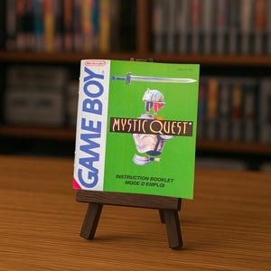 Peut inclure: Un livret d'instructions vert pour Game Boy, intitulé "Mystic Quest", est présenté sur un chevalet en bois miniature. Le livret présente une épée et une illustration de personnage. Le texte "GAME BOY" est imprimé verticalement sur le côté gauche.