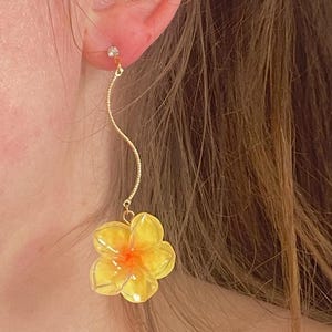 Może przedstawiać: Złoty kolczyk z małym diamentem i falistą zawieszką. Zawieszka kończy się żółtym kwiatem z pomarańczowym środkiem. Kolczyk jest noszony na uchu.