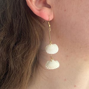 May include: Gold-tone drop earrings with two white seashell charms. The earrings have a hook fastening and a delicate design. The seashell pendants are intricately detailed, offering a natural, coastal look.