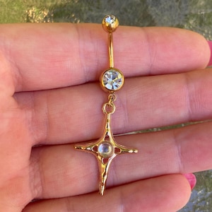 May include: A gold-toned navel ring featuring a curved barbell with a clear round crystal at the top and a larger clear crystal in the centre. A four-pointed star-shaped charm hangs from the bottom, set with a small iridescent round stone.