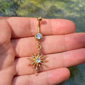 Puede incluir: Un piercing para el ombligo de color dorado con un dije de sol. El anillo presenta una pequeña gema transparente en la parte superior y una gema redonda más grande en la parte inferior. El dije de sol tiene múltiples rayos puntiagudos y una gema central.