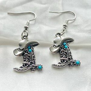 Pode incluir: Um par de brincos pendentes em tom prateado com formato de botas de cowboy. Cada bota apresenta detalhes de volutas, uma pequena espora no calcanhar e tr&ecirc;s pequenos detalhes redondos na cor turquesa no design.