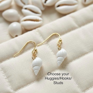 Puede incluir: Pendientes de concha blancos con ganchos dorados. Se muestran sobre una tela crema acolchada, con varias conchas marinas esparcidas. El texto dice "Choose your Huggies/Hooks/Studs".