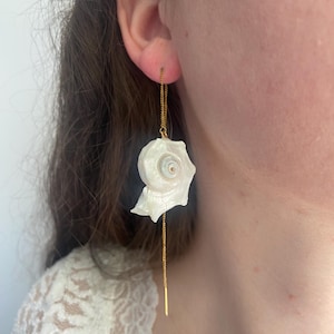 Peut inclure: Boucles d'oreilles coquillage. Chaque boucle d'oreille présente un coquillage blanc en spirale suspendu à une fine chaîne dorée. Les boucles d'oreilles sont conçues pour être portées à travers l'oreille, la chaîne s'étendant vers le bas.