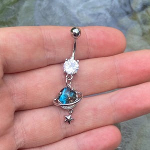 Könnte beinhalten: Ein silberner Bauchnabelpiercing mit einem klaren Edelstein, einem Planetenanhänger mit blauen und schwarzen Details und einem kleinen Sternanhänger. Der Ring hat ein gebogenes Stangendesign.