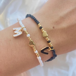 May include: Two beaded bracelets. One is white with a gold-coloured letter "M" charm and gold heart charms. The other is black with a gold-coloured letter "V" charm and gold heart charms. The bracelets are on a wrist.