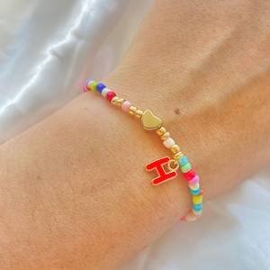 Puede incluir: Una pulsera de cuentas con un dije de corazón dorado y un dije de la letra "H" roja. La pulsera presenta una mezcla de cuentas coloridas. La pulsera se lleva en la muñeca.