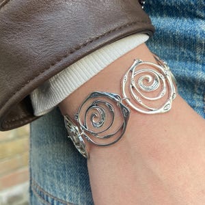 Könnte beinhalten: Ein silbernes Armband mit einem Spiraldesign. Das Armband besteht aus miteinander verbundenen kreisförmigen Formen mit einem durchbrochenen Muster. Der Schmuck wird am Handgelenk getragen, mit einer braunen Lederjacke und Jeans im Hintergrund.