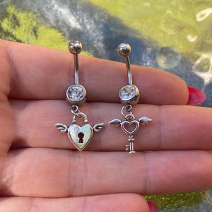 Peut inclure: Deux piercings de nombril argentés. Chaque bijou a une barre incurvée avec une gemme transparente en haut et en bas. L'un présente un cadenas en forme de cœur avec des ailes, l'autre une clé ailée.