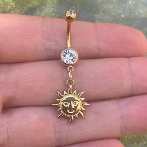 May include: A gold-colored belly button ring with a sun charm. The ring features a curved barbell with a clear gem at the top and a larger gem in the middle. The sun charm has a smiling face and radiating rays.