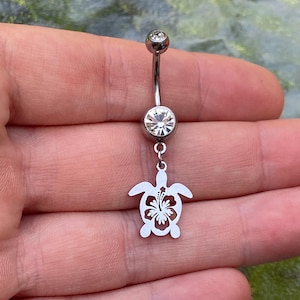 May include: A silver-toned curved barbell belly button ring featuring a small clear round gemstone at the top and a larger clear round gemstone in the centre. A dangling silver-toned charm shaped like a sea turtle with a hibiscus flower cutout.