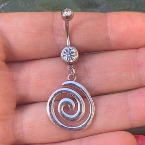 Puede incluir: Un piercing para el ombligo plateado con un diseño de espiral. El anillo presenta una barra curva con una gema redonda y transparente en la parte superior y una gema facetada más grande en el centro. Un dije de espiral plateado cuelga debajo.