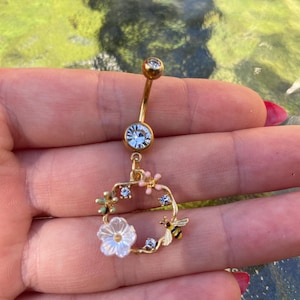 May include: Gold-coloured belly button ring with a floral design. The ring features a clear gem at the top and bottom, with a charm of a flower, bee, and small pink and blue flowers. The charm is a circular wreath.