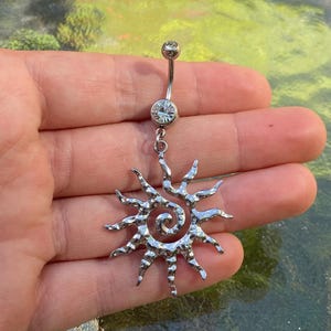 Puede incluir: Un piercing para el ombligo de color plateado con un diseño de sol. El anillo presenta una barra curva con una gema transparente en la parte superior e inferior, y un colgante de sol con un centro en espiral y rayos puntiagudos.
