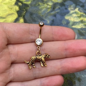 Peut inclure: Un piercing de nombril doré composé d'une barre incurvée avec un petit cristal transparent en haut et un cristal plus grand au centre. Une breloque en forme de léopard tacheté avec une finition texturée pend du cristal inférieur.