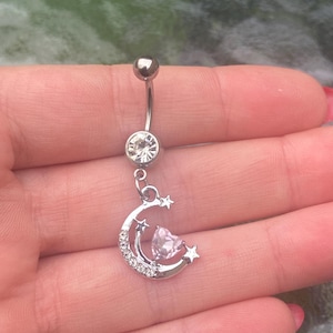 Può includere: Un piercing per ombelico argentato con una barra curva, una sfera superiore e una grande gemma trasparente. Il ciondolo presenta un design a mezzaluna con un cuore rosa e piccole stelle, impreziosito da cristalli trasparenti.