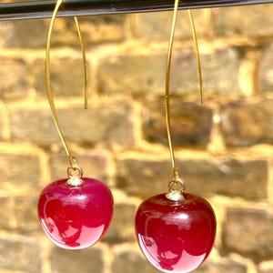 May include: A pair of drop earrings featuring two glossy, deep red cherry-shaped charms suspended from thin, curved gold-toned metal ear wires. The cherries have a smooth, reflective surface and are attached to the wires by small gold-coloured jump rings.