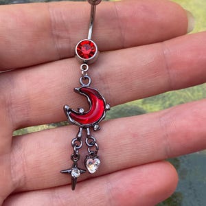 Puede incluir: Un piercing para el ombligo de barra curva plateada con un cristal rojo redondo en la parte superior. Un dije cuelga de la parte inferior, compuesto por una luna creciente roja, un pequeño corazón de cristal transparente y una estrella.