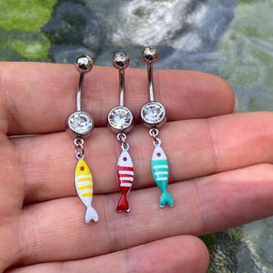 Könnte beinhalten: Drei silberfarbene, gebogene Bauchnabelpiercings, jeweils mit einem klaren, runden Kristall an der Basis und einem hängenden Fisch-Anhänger. Die Fisch-Anhänger sind gelb, rot und türkis emailliert und mit weißen Querstreifen versehen.