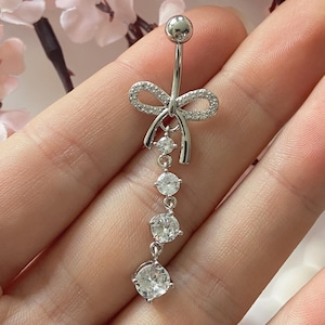 May include: A silver belly button ring featuring a bow design encrusted with small clear crystals. Below the bow, a series of four round, clear, sparkling crystals are suspended, creating a dangling effect. The top of the ring has a small, round silver ball.