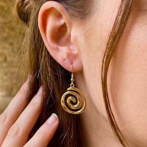 May include: A gold-toned metal dangle earring featuring a polished spiral design. The earring is suspended from a simple hook wire, showcasing a smooth, reflective surface that forms a circular, coiled pattern hanging from the earlobe.