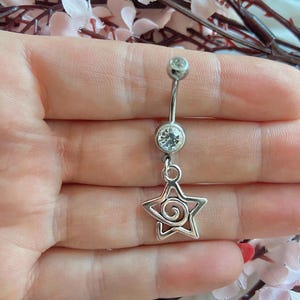 Puede incluir: Un piercing de ombligo plateado con un dije de estrella colgante. El anillo presenta una gema transparente en la parte superior y una gema más grande en el centro. El dije de estrella tiene un diseño en espiral.