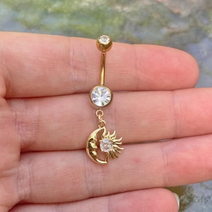 Könnte beinhalten: Goldfarbener Bauchnabelring mit Himmelsdesign. Die Oberseite und die Mitte weisen klare, runde Edelsteine auf. Ein Anhänger aus Mond und Sonne mit einem zentralen Edelstein hängt von unten. Der Schmuck wird in einer Hand gehalten.