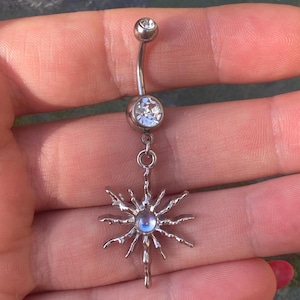 May include: A silver belly button ring with a curved barbell design. The top and bottom balls are clear, round gems. A sunburst charm with a central blue gem hangs from the bottom gem. The jewelry is displayed against a blurred background.