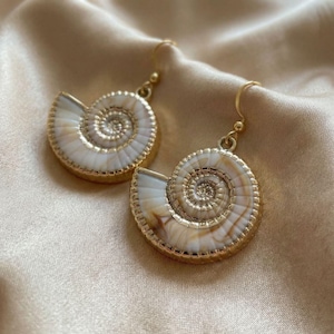 May include: A pair of drop earrings featuring white and tan spiral seashell charms. Each shell is encased in a thin gold-toned metal border with a matching gold-toned hook wire for attachment. The shells display natural swirling patterns.