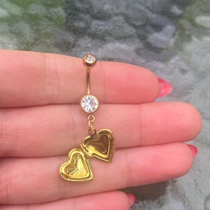 May include: Gold-colored belly button ring with a curved barbell design. The top and center feature clear, round gemstones. A heart-shaped locket with a butterfly design hangs from the center gem. The locket is open, revealing the interior.